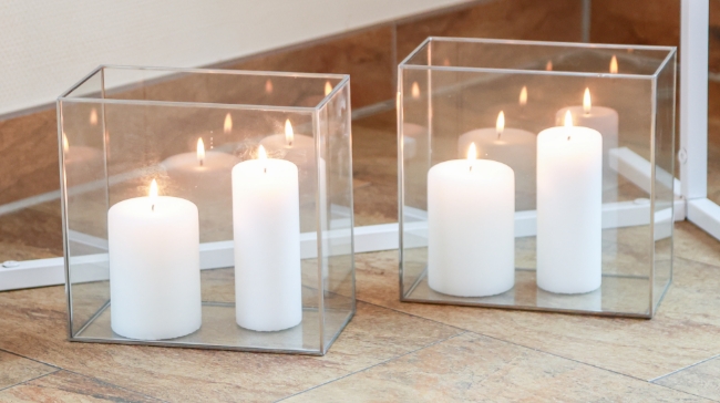 Two sets of white pillar candles burning inside clear square glass holders on a tiled floor reflecting in a mirror behind.