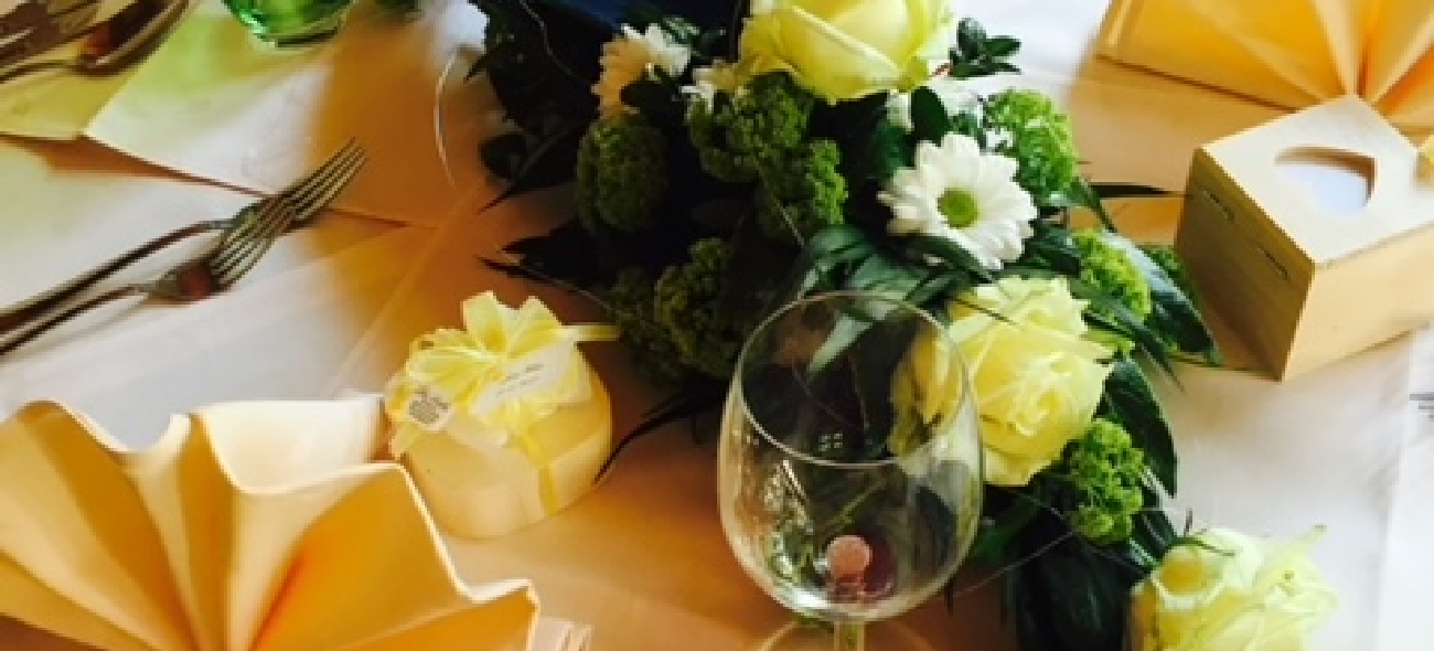 Table set with yellow folded napkins, a wine glass, cutlery, a small gift box with a yellow ribbon, and a floral centerpiece featuring yellow roses and white daisies.