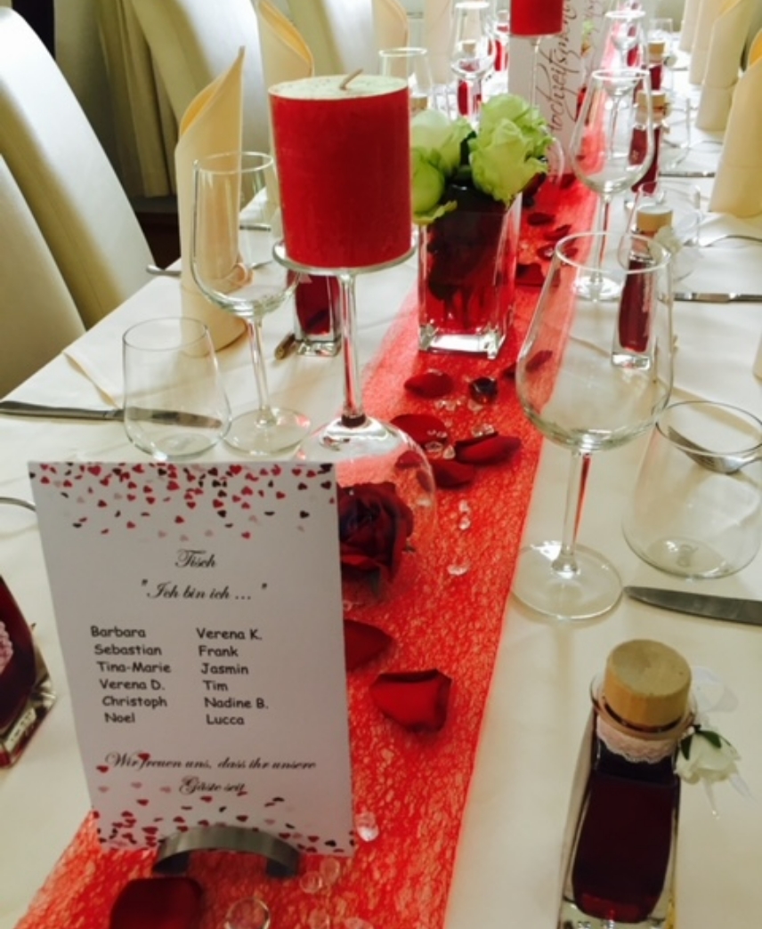Elegant table setting with white chairs, red runner, red candles, glassware, rose petals, and a seating card listing guest names in German.